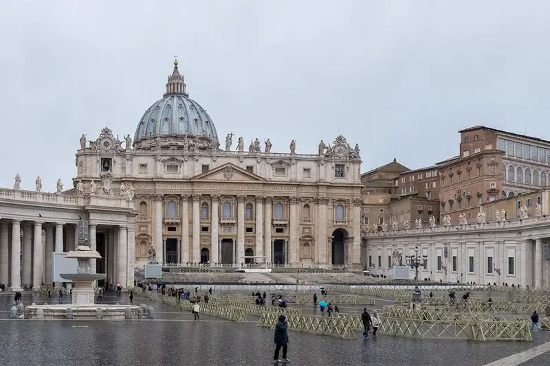 St. Peter's Basilica in Vatican City on a cloudy day.	People walk through the large, wet plaza, partially cordoned off with temporary barriers.
