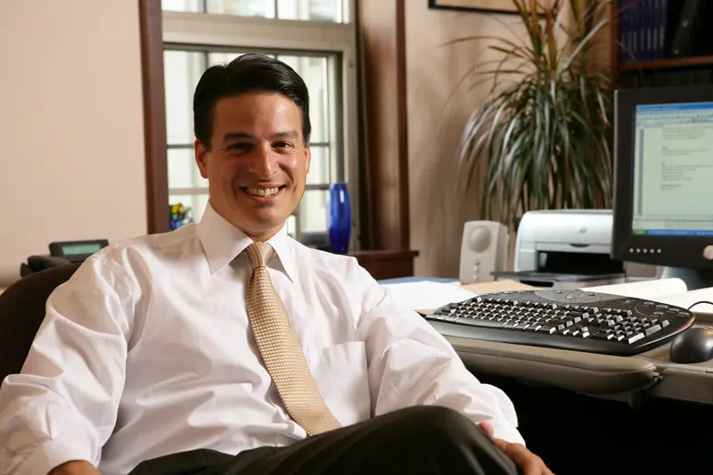Man in a white shirt and gold tie smiles while sitting at his desk in an office.