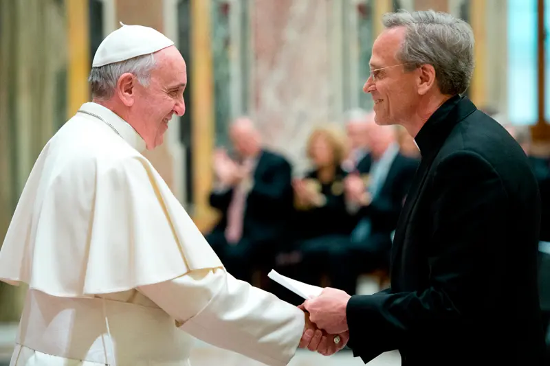 Pope Francis, wearing white papal vestments, smiles and shakes hands with a man in a dark suit and clerical collar, while also handing him a small white booklet.	A blurred audience is visible in the background.