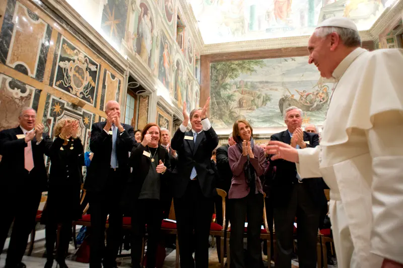 Pope Francis smiles and gestures towards a group of people applauding him in a frescoed hall.