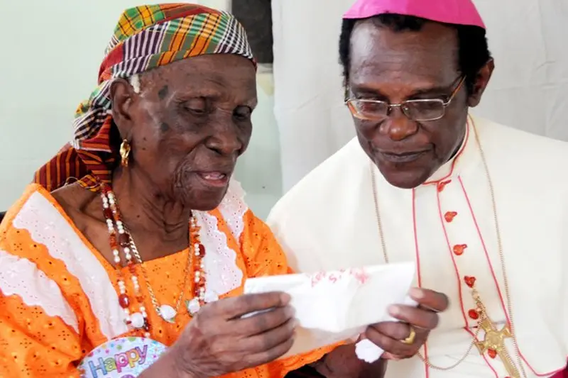An elderly woman wearing a colorful headwrap and orange dress looks at a card held by a person wearing a pink Bishop's mitre and white vestments.