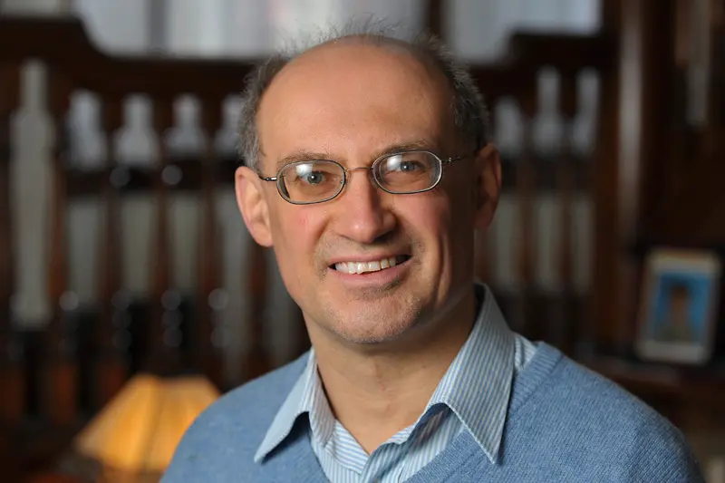 Headshot of a man with glasses, wearing a light blue sweater over a light blue and white striped collared shirt, smiling at the camera.	A blurry background shows wooden furniture and a lamp.