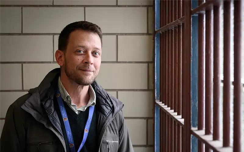 A white man wearing a gray winter jacket and a lanyard is standing next to prison bars.