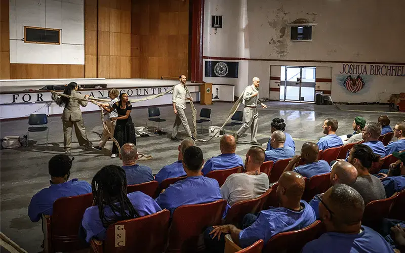 Five actors holding a long strip of fabric between them stand in front of a seated crowd of prisoners. They are performing one of Shakespeares plays.