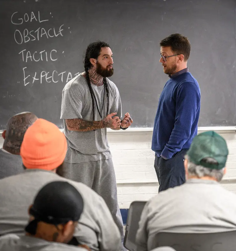 A man in gray sweats, with tattoos and braided hair, speaks with clasped hands to another man and a classroom of incarcerated individuals.