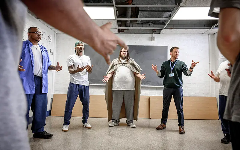 In a classroom setting, incarcerated individuals participate in a drama class. One person, wearing a makeshift cape, stands center stage with arms outstretched, seemingly portraying a character. Flanking them are two other incarcerated individuals, engaged in expressive gestures. A drama instructor guides the group, his hands raised in a directorial manner.