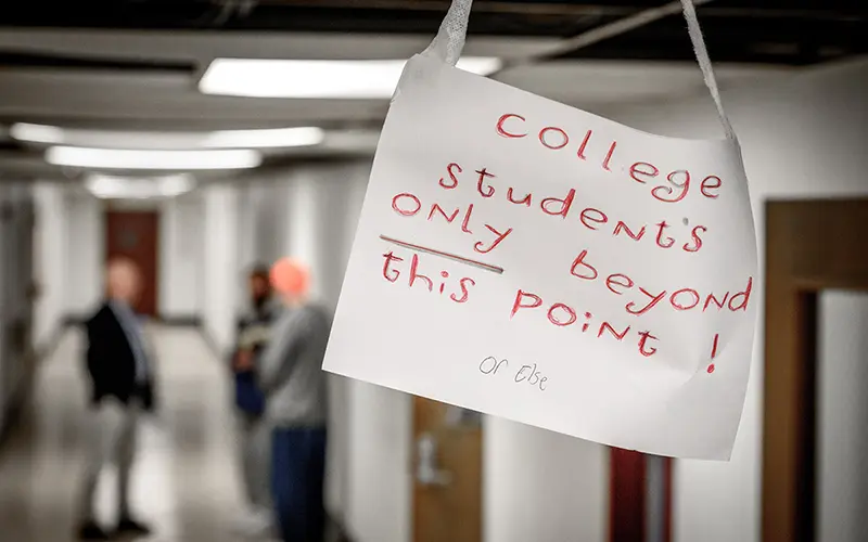 A handwritten sign on white paper reads, College students only beyond this point! with or else written smaller beneath. The sign hangs in a hallway with fluorescent lighting.