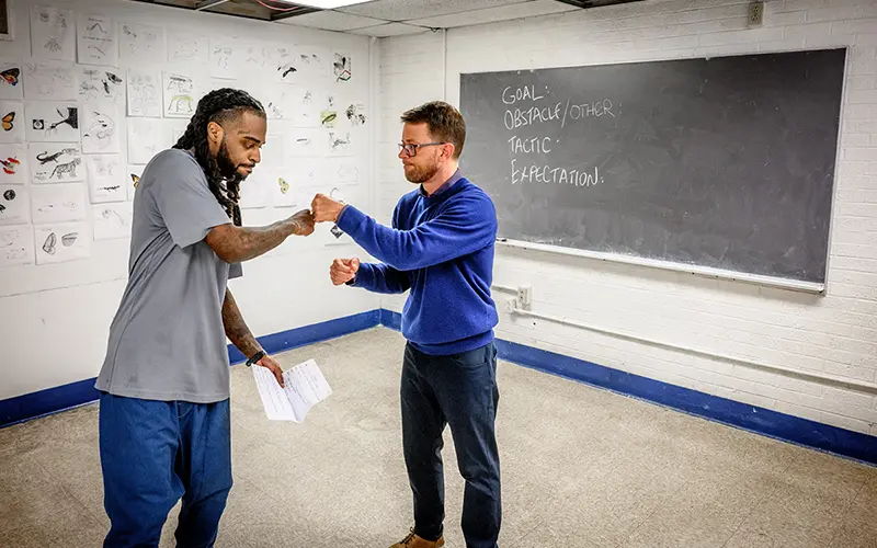 Two men perform a short dramatic scene in front of a blackboard in a classroom. One man wears gray and blue athletic clothing and holds a sheet of paper. The other man wears a blue sweater and khakis.