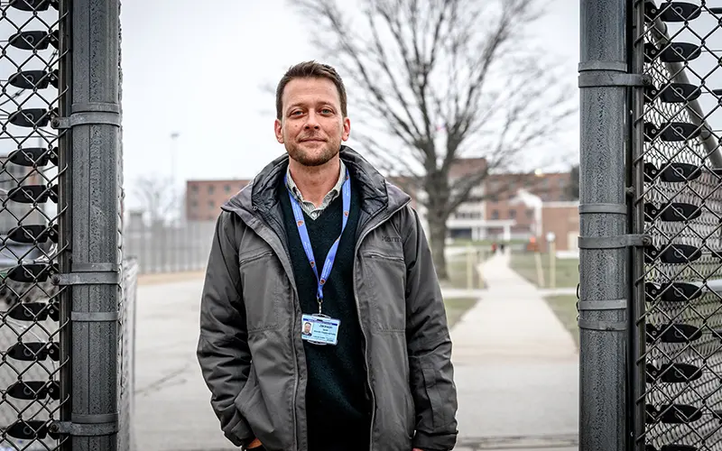 A white man in a gray winter coat stands in between the gates of the prison entrance.