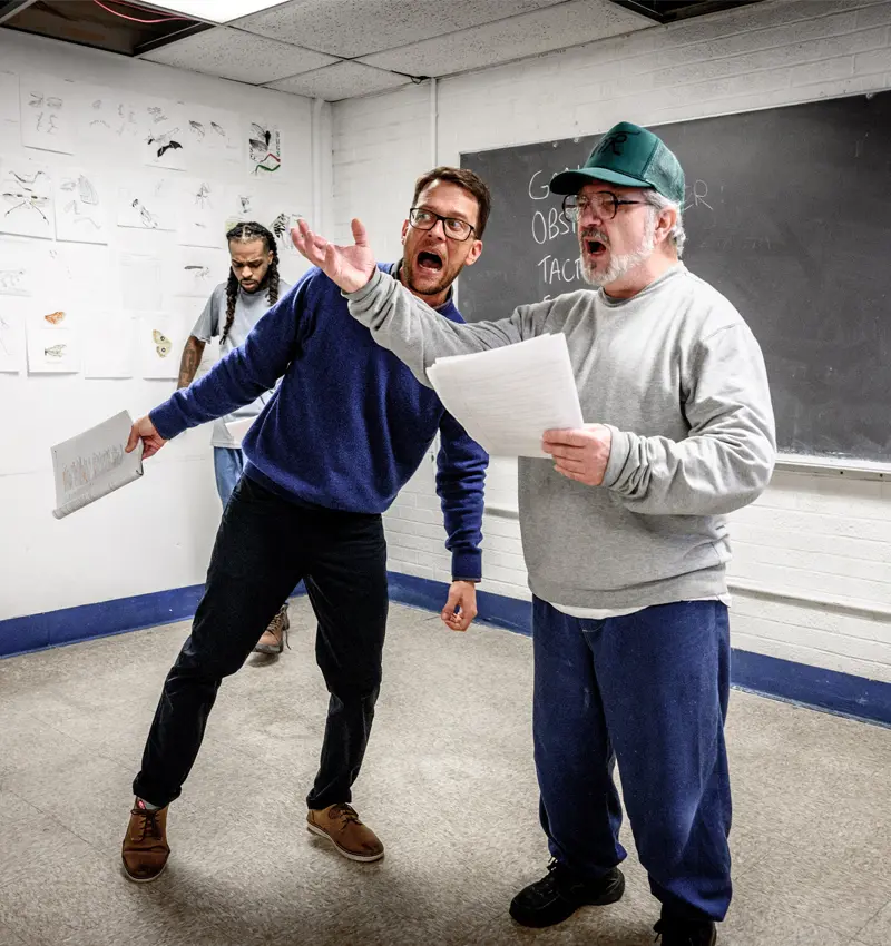 A white man stands at the front of a classroom with one arm raised has a script in the other hand. Next to him, the teacher is acting along and making a dramatic face.