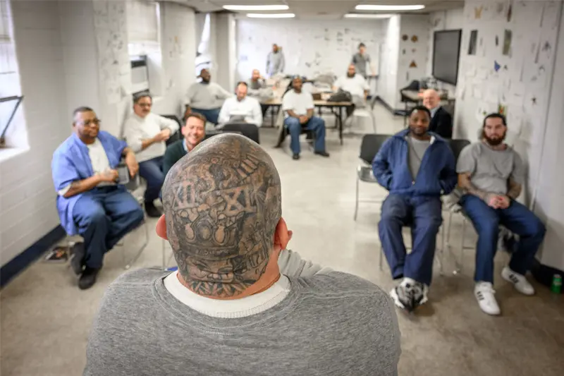The back of a tattooed bald head as it looks into a classroom of men.