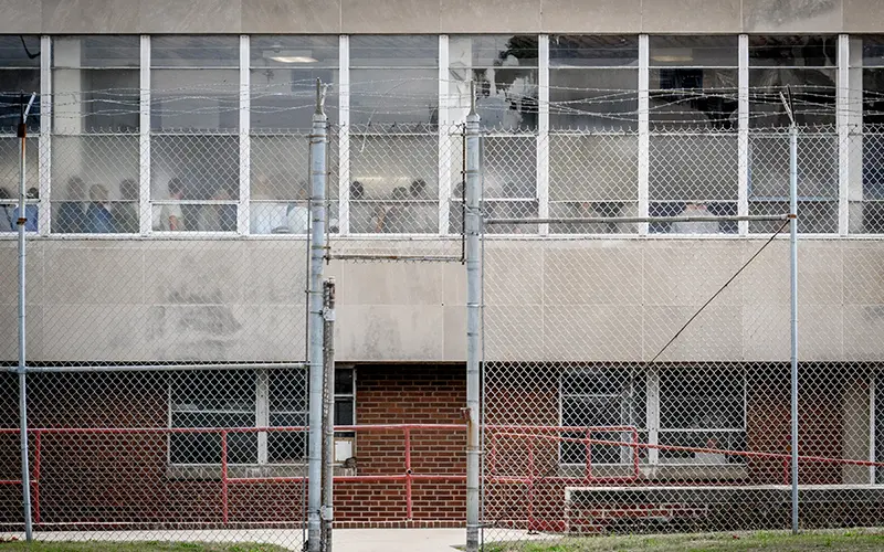 A chain-link fence with barbed wire atop it surrounds a non-descript building. Through the upper story windows, blurred figures can be seen inside. A red railing and gate lead to a brick portion of the building at ground level.