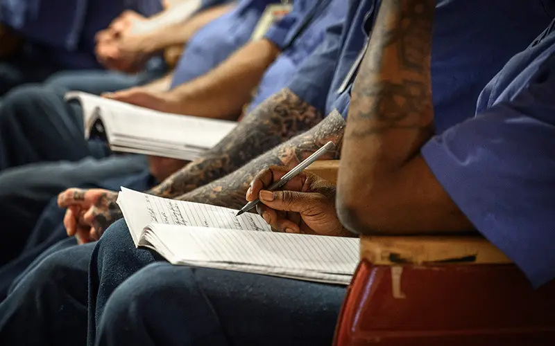 lose-up of several individuals wearing blue attire, likely incarcerated individuals, sitting in rows. Focus is on the hands of one person writing in a notebook with a pen. Their arms are adorned with tattoos.	Others in the row also appear to be taking notes in a classroom or meeting setting.