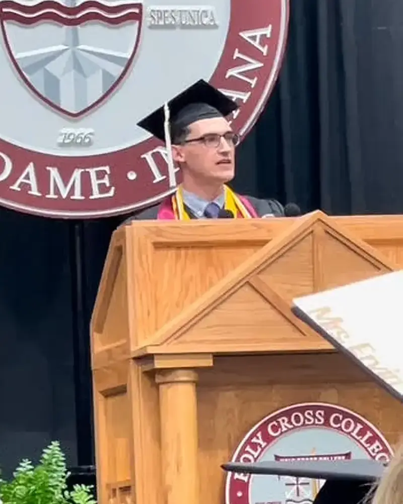 A white male stands at a podium in his graduation cap and gown.