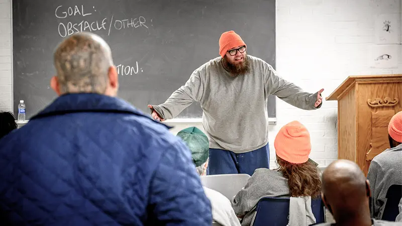 A white man with a beard and wearing a beanie is standing at the front of a classroom with his arms extended out to his sides.