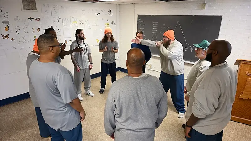 A group of incarcerated individuals stand in a circle in a classroom. Some wear orange and gray beanies. A chalkboard with writing and a graph is behind them. Drawings and photos of butterflies and insects are taped to the wall. They appear to be engaged in a group activity.