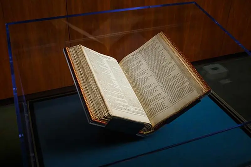 An antique, open book rests on a clear display stand within a glass case. The book's aged, yellowed pages are filled with small, densely printed text. A dark, possibly leather, binding protects the delicate pages. The display case appears to be situated in a dimly lit room with wood paneling in the background.
