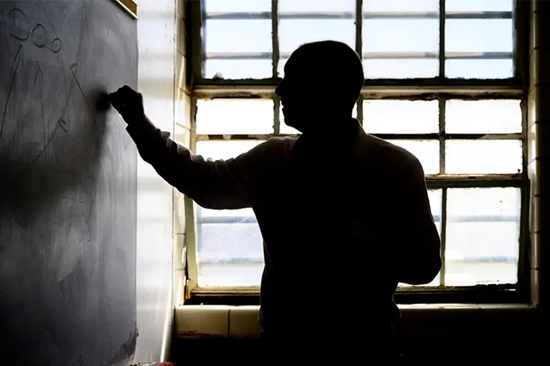 A man stands in profile writing on a chalkboard. He is silhouetted against a bright window.