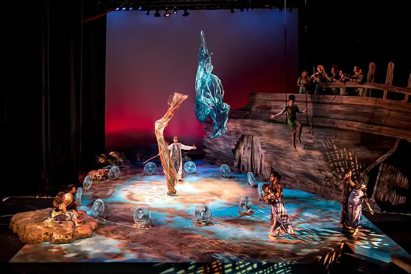 A vibrant theater scene depicts a performance. A figure in green is suspended in the air near a wooden ship's hull. Other actors are positioned across the stage, some on a rocky outcrop, while others stand on a painted seascape.  A central figure with outstretched arms stands near a swirling teal cloth suspended mid-air. Transparent globes encircle the actors, adding to the magical ambiance.