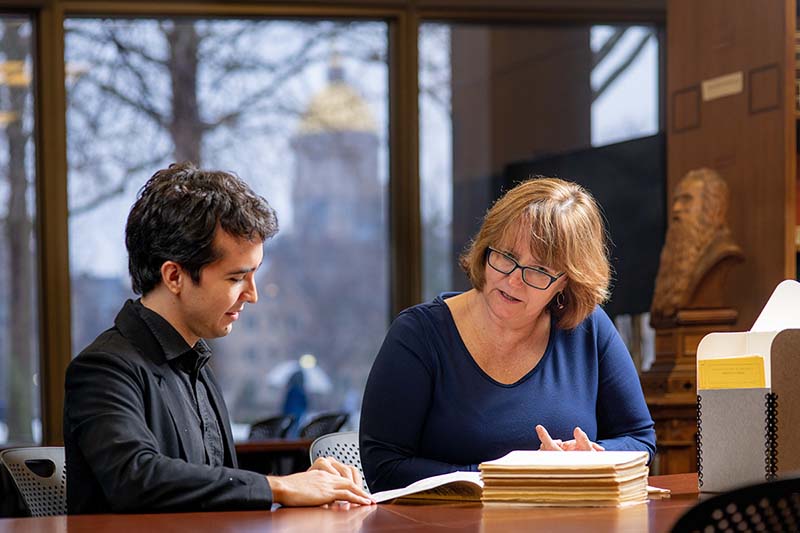 A man and a woman sit at a table in the library with the stacks of journals. The golden dome of the Main Building is visible out the window behind them.