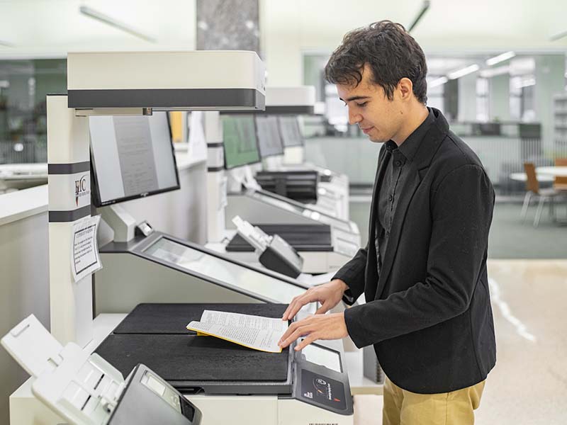 The student stands at a scanner in the library with the journal under the scanner.