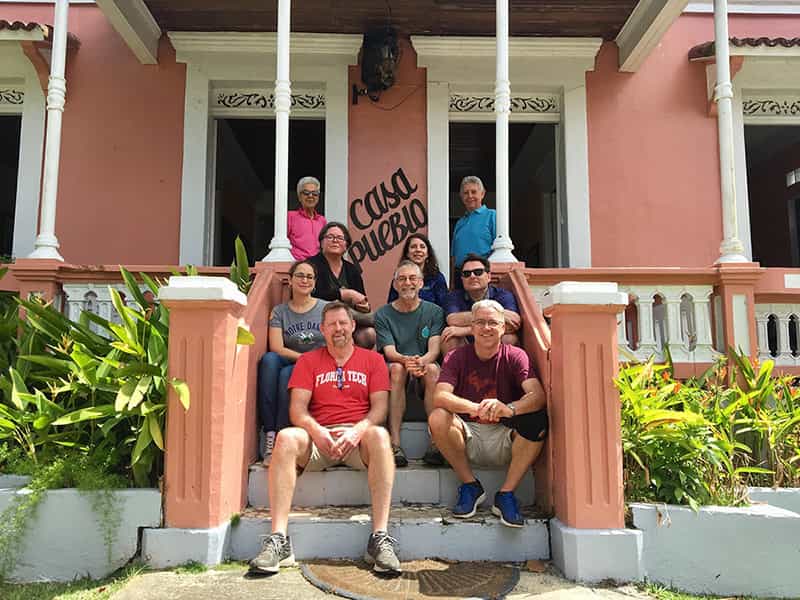 A group of people sit on steps in front of a home.