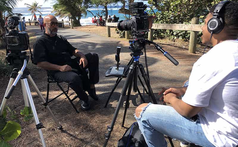 A student sits behind a camera interviewing a man outside near the ocean.
