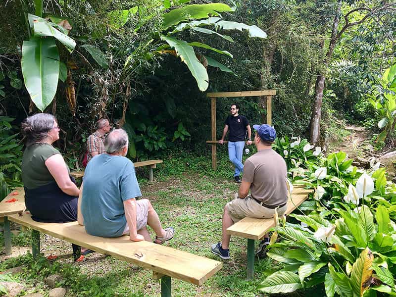 Four people sit on wooden benches and listen to another who leans against a railing.