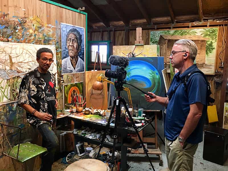 A man stands in front of artwork while another films an interview.