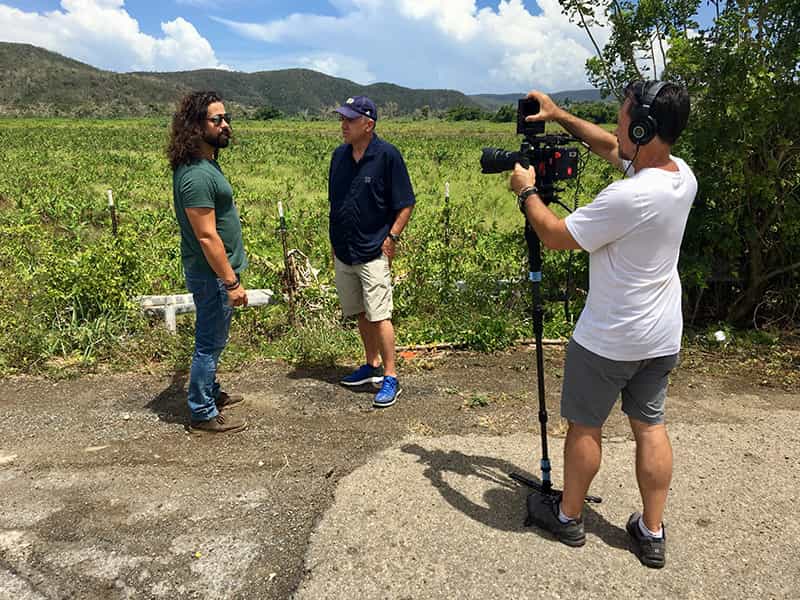 A man interviews another standing near an open field as a videographer films their interview.