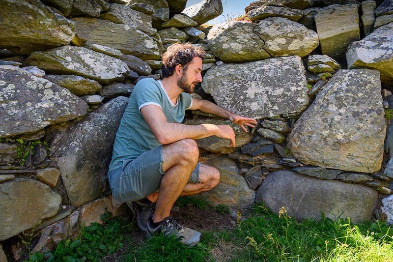 A man crouches next to a stone wall, with multiple crosses carved into stone.
