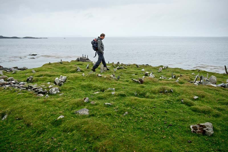 Ian Kuijt walks through a stoney cemetary beside the ocean. Green grasses, cloudy day.
