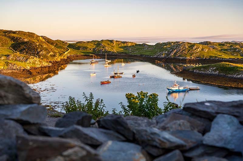 A small harbor with several boats in it during a golden sunset. Small green rocky hills surround the harbor.