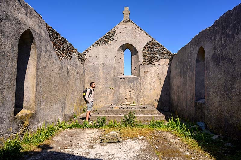 A man stands inside an ancient monastic building made of stone. The ceiling is gone, open to a blue sky. Tall grasses grow inside along the periphery.