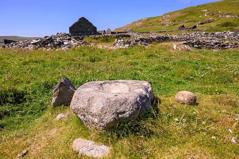 A large stone with a circular depression on top, set against a backdrop of green grass and the ancient monastery.
