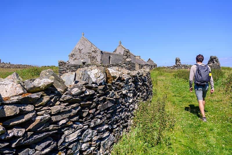 A man walks along a stone wall, with medieval structures in the background. Green grasses and blue skies.