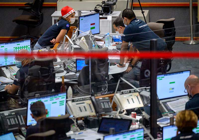 A female at a desk, wearing a red Santa hat, leans forward to talk to two	males looking at a computer. They talk amongst themselves. Others sit at computers in the foreground which is out of focus.