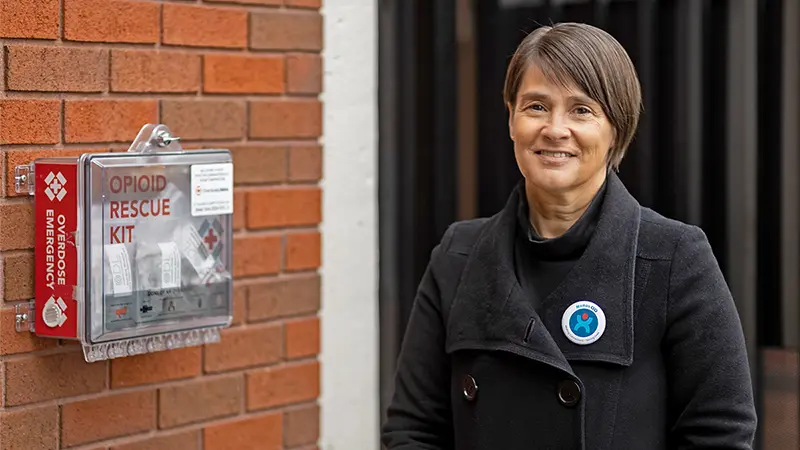 Joanne Kelley Codgell stands next to an opioid rescue kit attached to a brick wall.