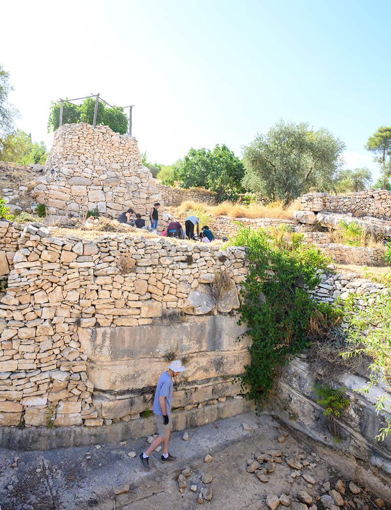 A group of students dig on top of a stone hill, their professor is below them on a lower level of the hill. There's rocks scattered everywhere and green trees in the foreground and background.