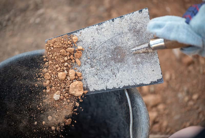 A close up shot ofsomeone using a trowel to transfer dirt to a bucket.