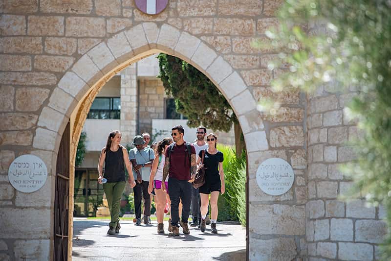 A group of students walk towards the camera, through a stone arch. A blurred out tree branch is in the foreground.