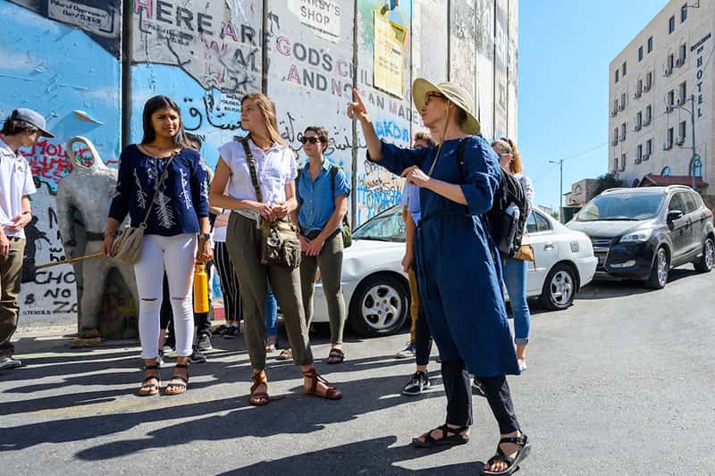 A women talking to a group of students about a colorful wall with graffiti on it.