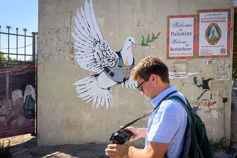 A student looking at his camera stands in front of a wall painting of a dove with an olive branch wearing a flak jacket and a target painted over its breast.