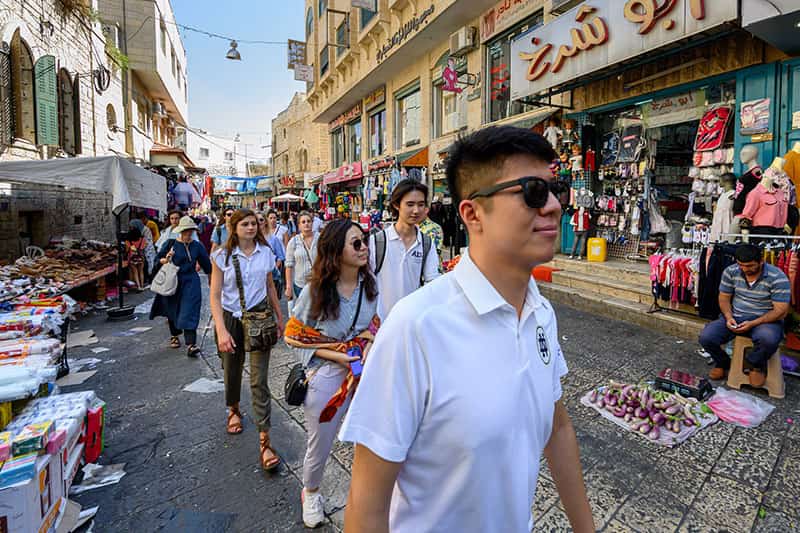 Students walk through a busy market. There are people sitting and standing on the side of the street, selling clothing, food, and other items.