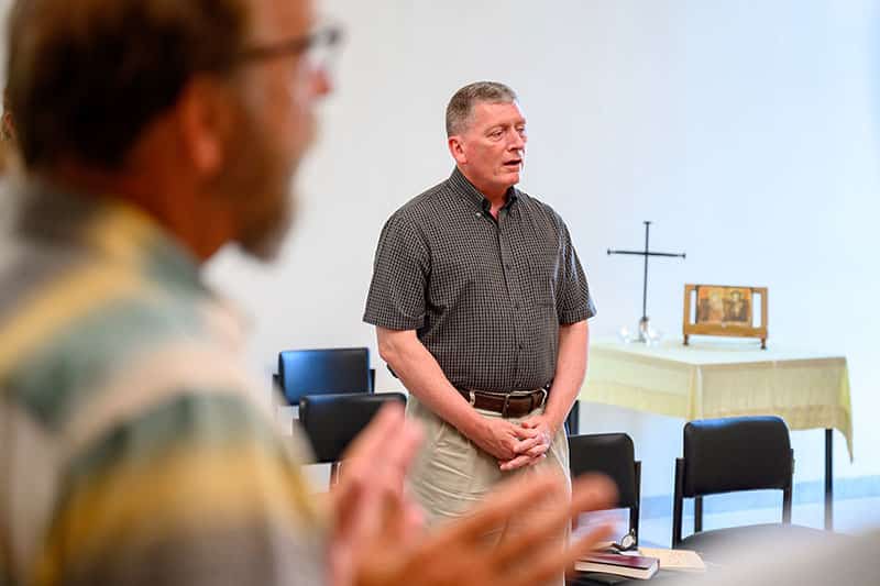 The camera focuses on a man standing with his fingers interlaced singing in a room, another man is blurred out in the foreground.