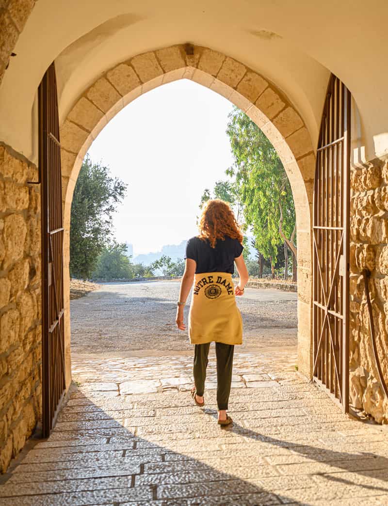 A student wearing a Notre Dame sweatershirt around her waist walks through a gate surrounded by bricks as the sun shines through the opening.