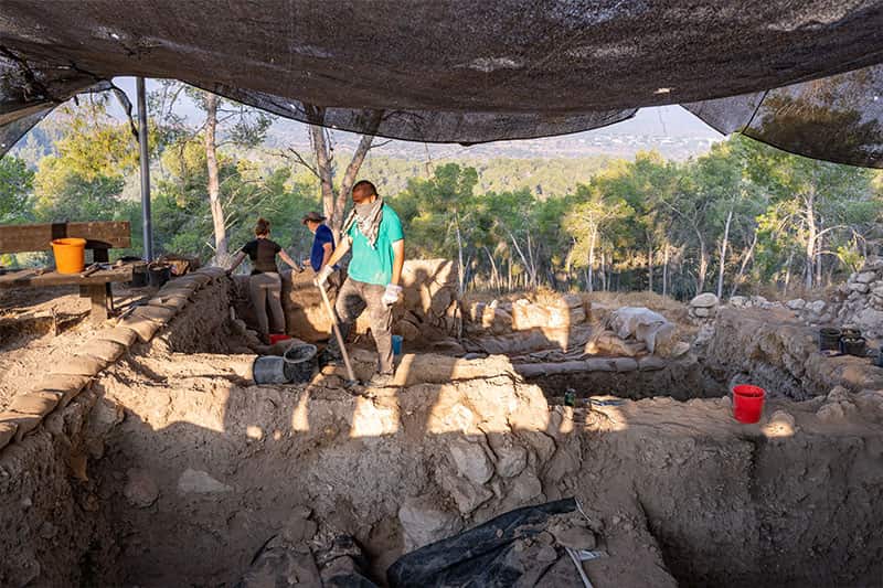 Three people work on excavations under a shelter made of fabric, one person wears a scarf around their face to protect them from the dust and dirt.