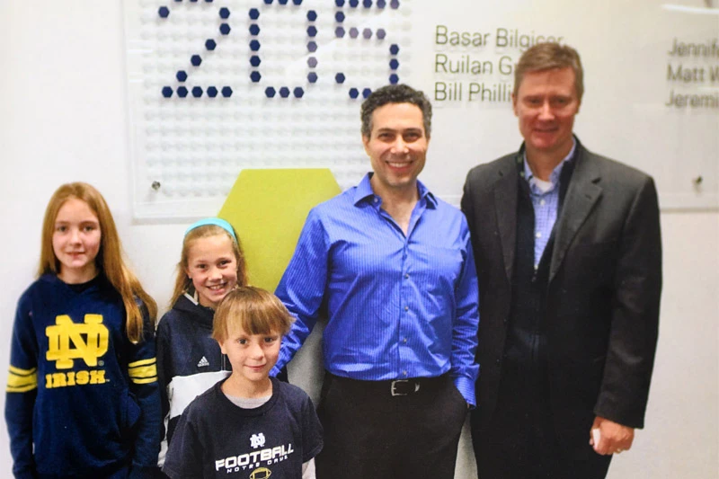 Two men and three children pose for a photo in front of a wall displaying names and a large '2015' created with small hexagons. The children wear Notre Dame apparel. One man wears a blue, pinstriped dress shirt and the other a dark suit jacket.