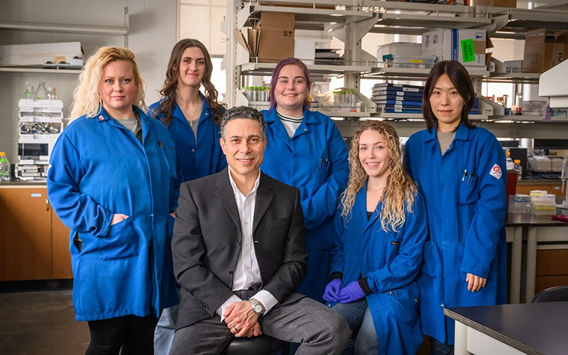 Six individuals, five in blue lab coats and one in a dark suit, pose for a photo in a laboratory setting.