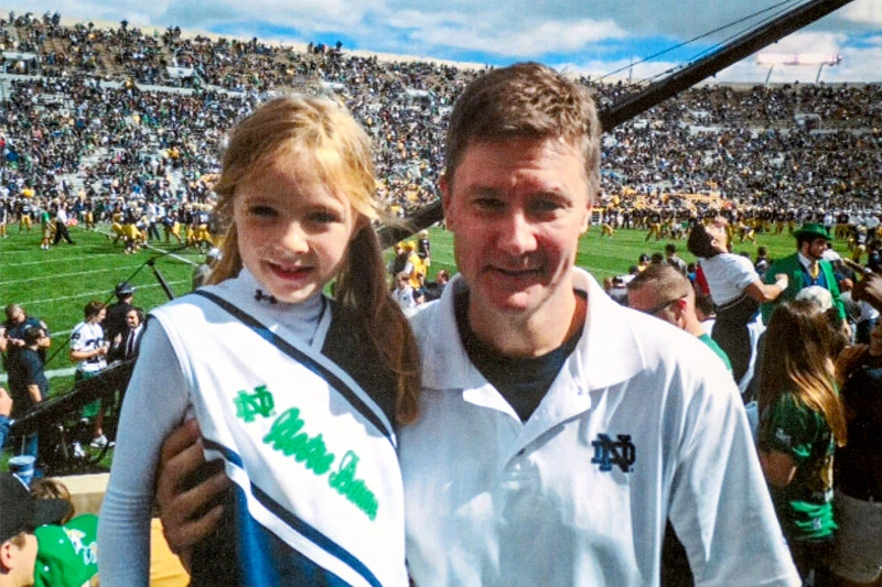 A smiling girl in a Notre Dame cheerleading outfit poses with a man in a white Notre Dame polo shirt in Notre Dame Stadium during a game.  The crowded stands and football field are visible in the background.